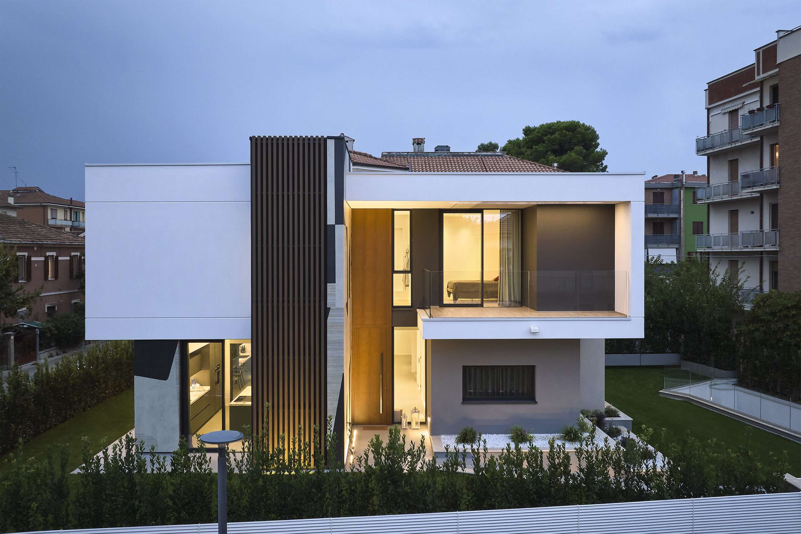 Villa con volumi bianchi e legno, con le aperture illuminate al crepuscolo